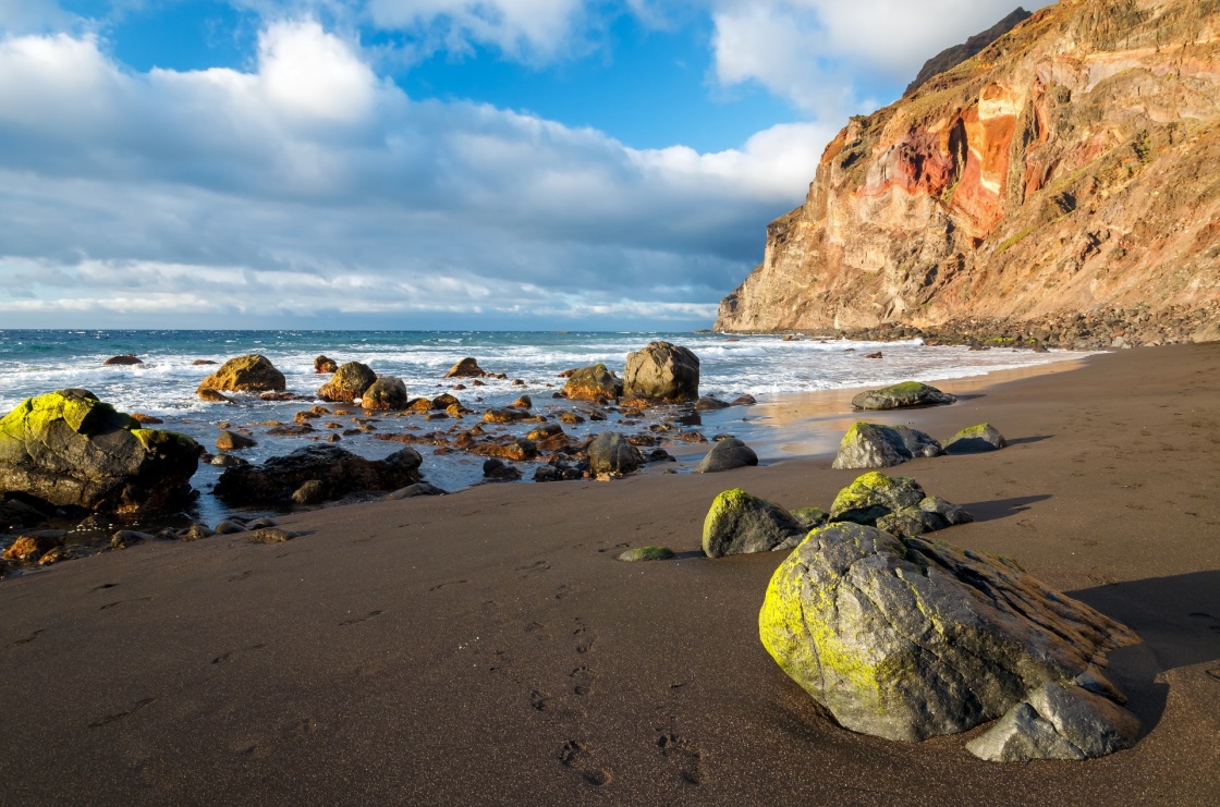 Rocks stones on volcanic black sand beach with white clouds on blue sky, Playa Ingles, La Gomera island